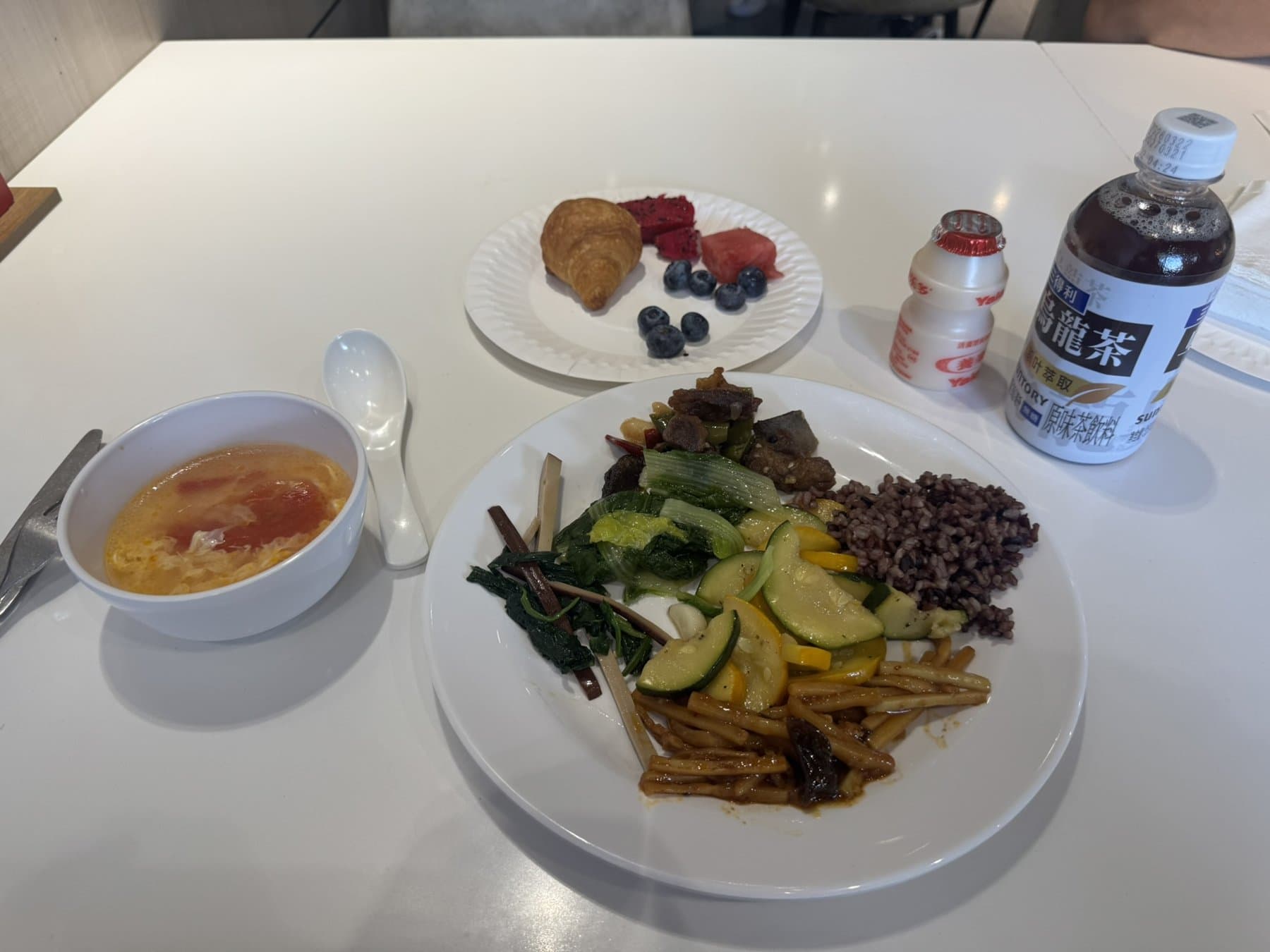 A factory canteen lunch tray: stir-fried vegetables, purple rice, egg-drop soup, a croissant with fruit, a Yakult and a bottle of oolong tea