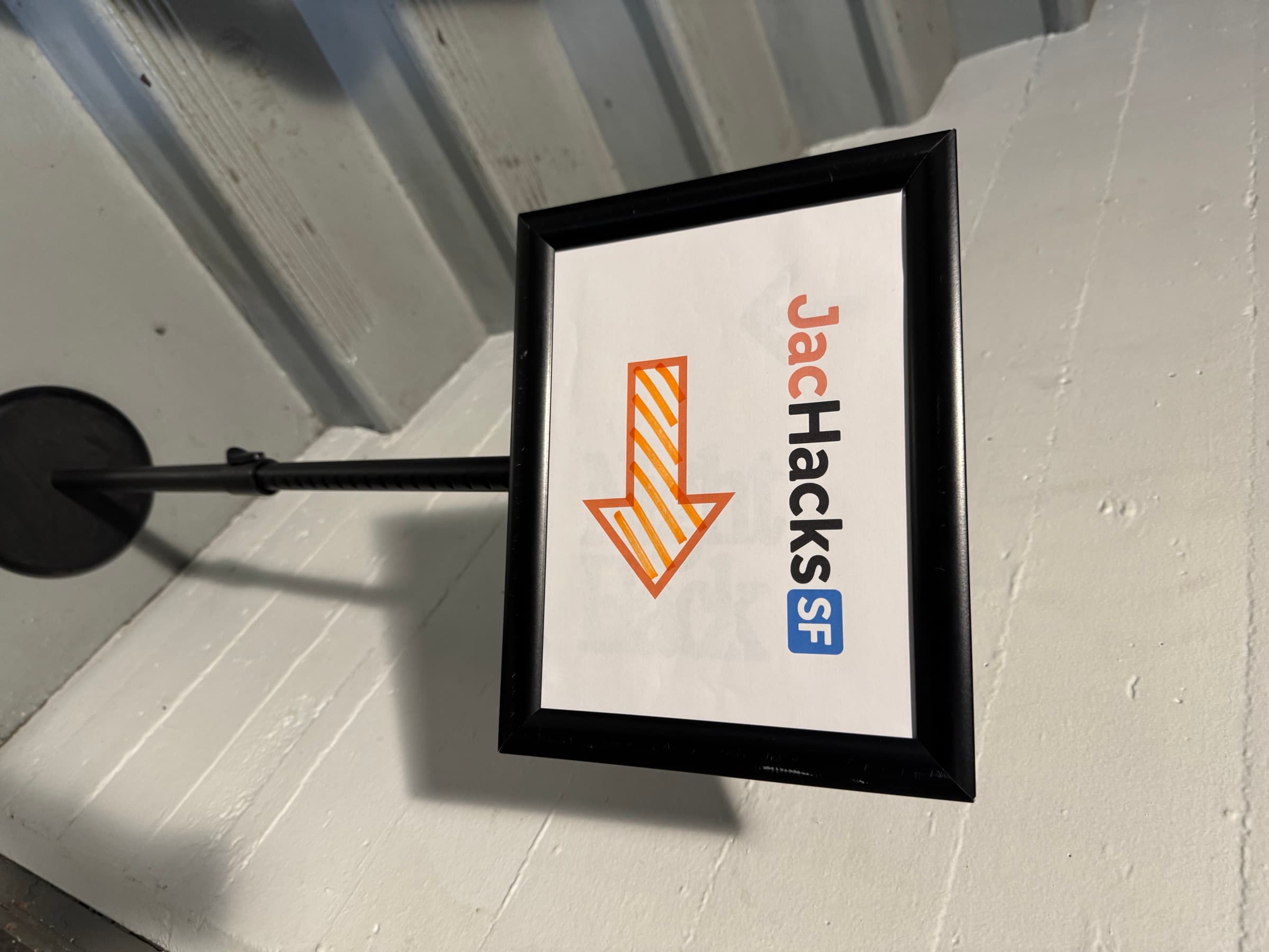 A framed JacHacks SF sign with an orange arrow, on a stand at the top of a stairwell